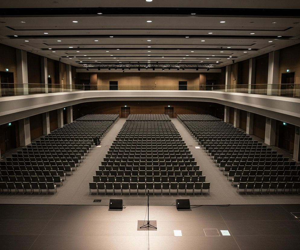 Przestronna, pusta aula lub sala konferencyjna o nowoczesnym wystroju. W centralnej części widać liczne rzędy ciemnoszarych krzeseł, ustawionych w trzech blokach przed sceną, oddzielonych dwoma szerokimi przejściami. Scena, w dolnej części kadru, jest oświetlona, na niej stoi statyw mikrofonowy oraz głośnik sceniczny. Nad krzesłami i wokół sali rozciągają się balkony z szklanymi barierkami i drewnianymi panelami na ścianach, a na suficie widoczne są liczne punkty świetlne.