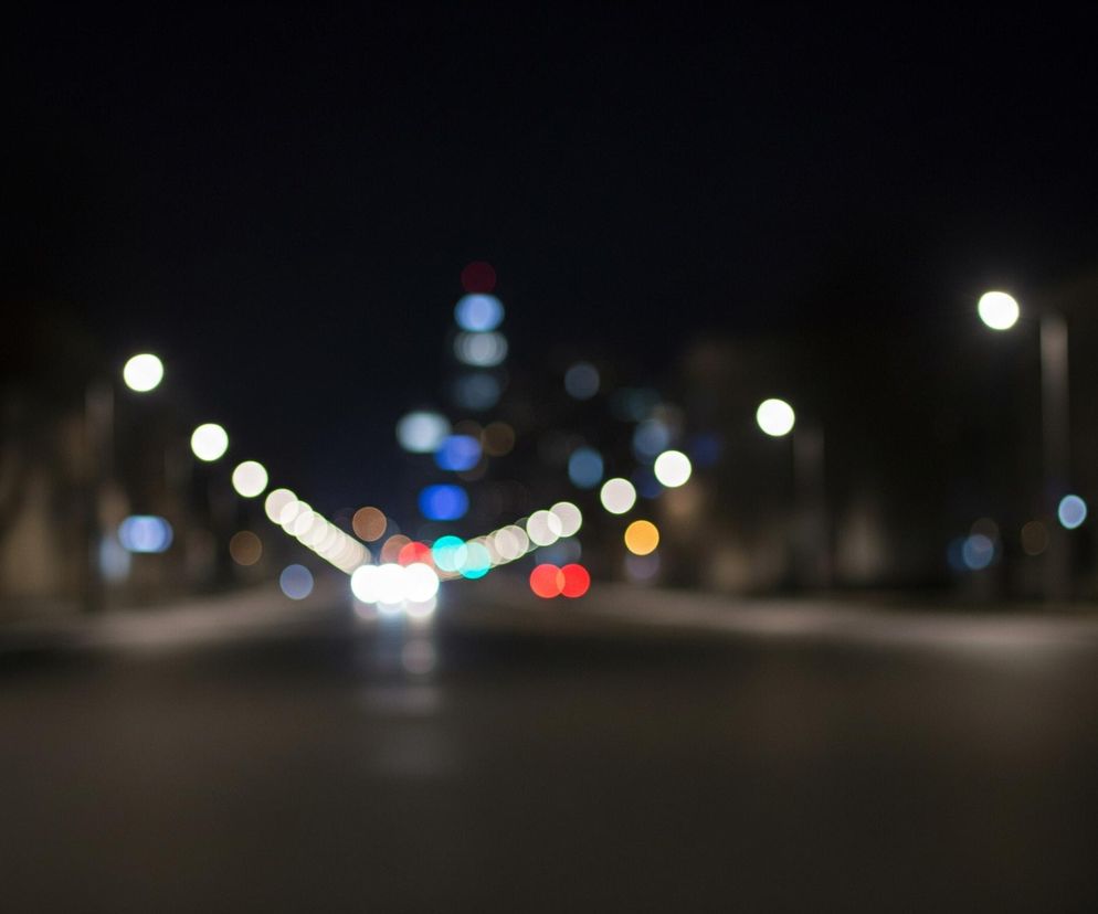 Na zdjęciu widoczna jest rozmyta nocna panorama miasta, z czarnym niebem zajmującym górną część kadru. Ulicę o ciemnoszarym odcieniu przecina pasmo świateł, głównie białych i żółtych, tworzących efekt bokeh. Po obu stronach drogi rozmieszczone są uliczne latarnie, również widoczne jako rozmyte, jasne punkty. W oddali, w centrum kadru, można dostrzec skupisko różnobarwnych świateł, w tym niebieskich, czerwonych i zielonych, przypominających oświetlenie wysokich budynków lub sygnalizację świetlną. Po prawej stronie zdjęcia zarysowuje się ciemna sylwetka budynku, a po lewej stronie znajdują się niewyraźne, ciemne obiekty przypominające drzewa lub inne zabudowania.