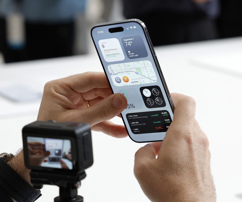Mężczyzna trzyma w dłoni najnowszy model iPhone'a z aktywnymi widżetami, w tym mapą, pogodą i notowaniami giełdowymi. Na pierwszym planie widoczna jest kompaktowa kamera nagrywająca ekran smartfona. Artykuł na Super Biznes analizuje, jak banki kuszą klientów, oferując w zamian za założenie konta nowe produkty Apple, jak ten prezentowany na zdjęciu.