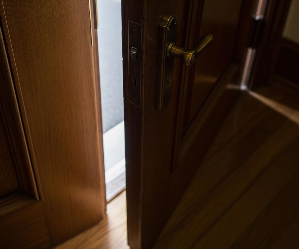 Drzwi z jasnego drewna, lekko uchylone, ukazują fragment wnętrza. Po prawej stronie widoczny jest mosiężny uchwyt drzwiowy z zamkiem, a w szczelinie między framugą a drzwiami prześwituje jasne światło. Podłoga z drewnianych paneli lub desek rozciąga się od dołu kadru, z wyraźnym światłem padającym na nią od lewej strony.