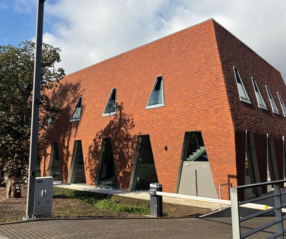 Nowoczesny budynek Ośrodka Szkoleniowo-Naukowego Okręgowej Izby Lekarskiej w Gdańsku z czerwoną elewacją z cegły i dachówek, oraz charakterystycznymi, trójkątnymi oknami, widoczny od strony podjazdu. O rozwiązaniach architektonicznych przeczytasz na Architektura Murator Plus.