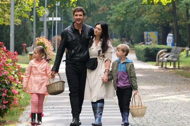 Adriana Kalska i Mikołaj Roznerski jako Iza i Marcin Chodakowscy z serialowymi dziećmi Mają (Laura Jankowska) i Szymkiem (Staś Szczypiński) spacerują w parku, trzymając koszyki. Można o tym przeczytać na naszym portalu.