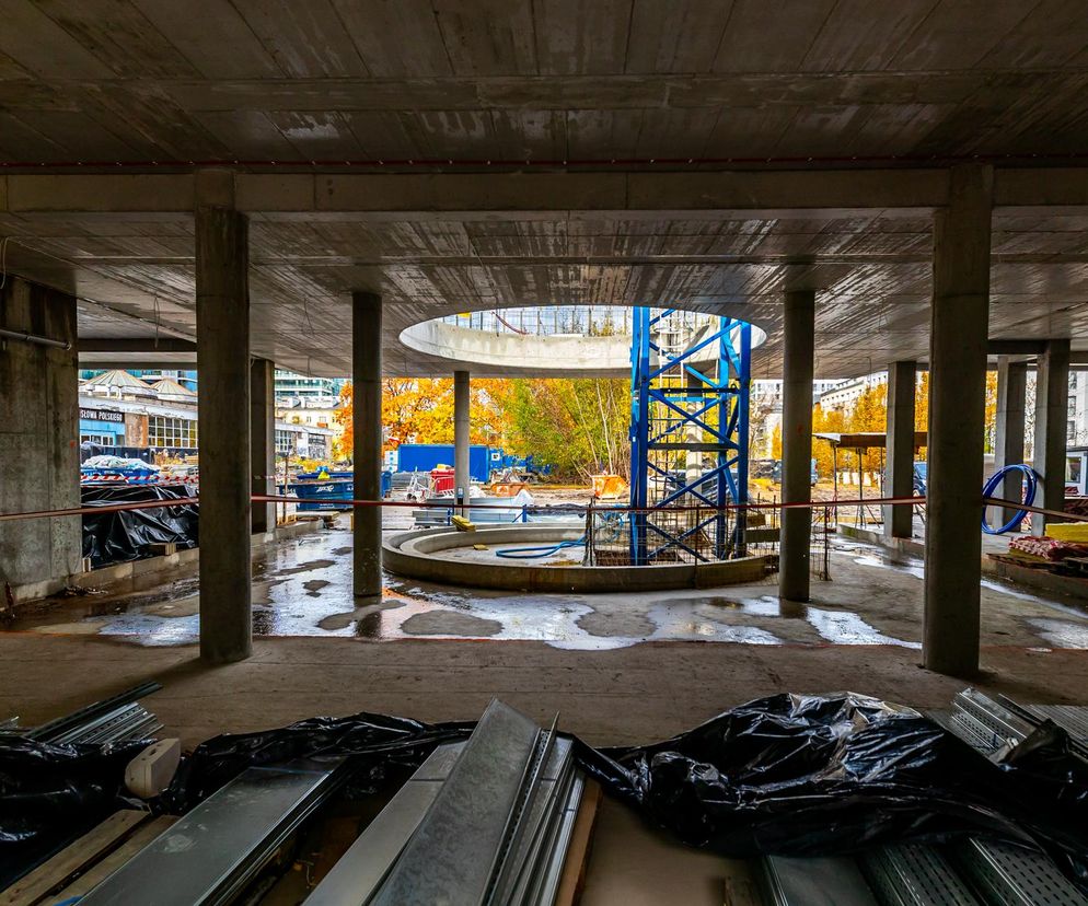 Plac budowy z niedokończonymi betonowymi konstrukcjami apartamentowca premium, widok z wnętrza na koparkę i jesienne drzewa. Obraz symbolizuje trwające inwestycje w luksusowe nieruchomości, o których więcej przeczytasz na Super Biznes.