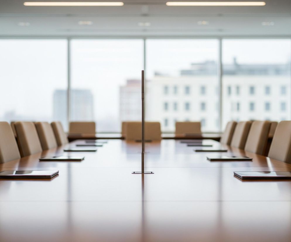 Centralnie położony jest długi, polerowany, drewniany stół konferencyjny w kształcie prostokąta, rozciągający się od pierwszego planu do oddali, po obu stronach którego stoją beżowe krzesła biurowe z wysokimi oparciami. Na stole leżą płaskie, ciemne urządzenia elektroniczne, prawdopodobnie tablety, w równych odstępach przed każdym krzesłem, tworząc odbicia na błyszczącej powierzchni. W połowie długości stołu pośrodku wystaje pionowy, smukły, ciemny element, prawdopodobnie rozdzielacz lub stojak.

W tle widoczne jest duże okno z jasnym, rozproszonym światłem wpadającym do pomieszczenia, przez które zarysowują się niewyraźne kształty budynków na zewnątrz. Pomieszczenie ma szare ściany i sufity, a pod sufitem widoczne są podłużne, świetlówki emitujące ciepłe światło.