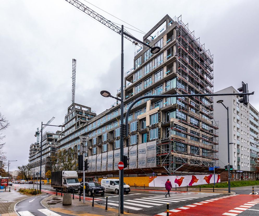 Nowoczesny budynek wielorodzinny przy ul. Anielewicza w Warszawie w trakcie budowy, z rusztowaniami i żurawiami, obok ruchliwa ulica i istniejące bloki. W tle jesienne drzewa. Więcej o kontrowersyjnej inwestycji przeczytasz na Architektura Murator Plus.