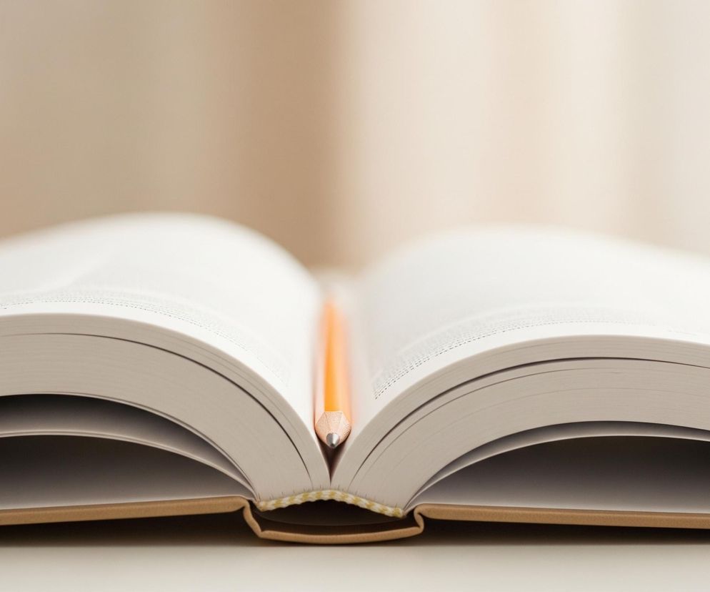 Otwarta książka leży na jasnej powierzchni, zajmując centralną część kadru. Pomiędzy jej stronami, w zgięciu, znajduje się żółty ołówek z widocznym grafitowym końcem. Kartki książki są jasne, niemal białe, z widocznym tekstem w górnych partiach, a twarda okładka ma kolor brązowy. Tło jest rozmyte, w odcieniach beżu i bieli, tworząc efekt miękkiego światła.