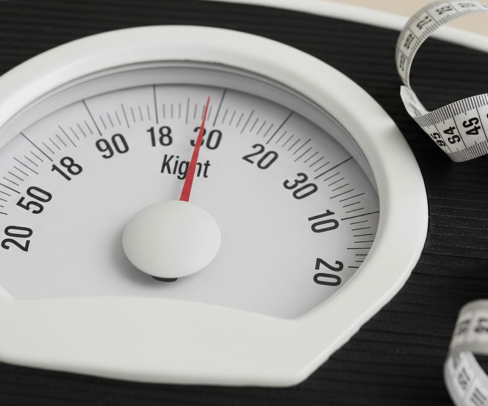 Centralnie umieszczona jest waga łazienkowa z okrągłym, białym cyferblatem, na którym widoczne są czarne cyfry i kreski, a także czerwona wskazówka skierowana w prawo na wartość około '25'. Pod cyframi '18 30' widnieje czarny napis Kight, a pod nim znajduje się białe, kuliste pokrętło. Tło wagi jest czarne z poziomymi, wąskimi żłobieniami. Po prawej stronie wagi leży biała miarka krawiecka, zwinięta w luźne kształty, z widocznymi na niej czarnymi numerami i podziałką.