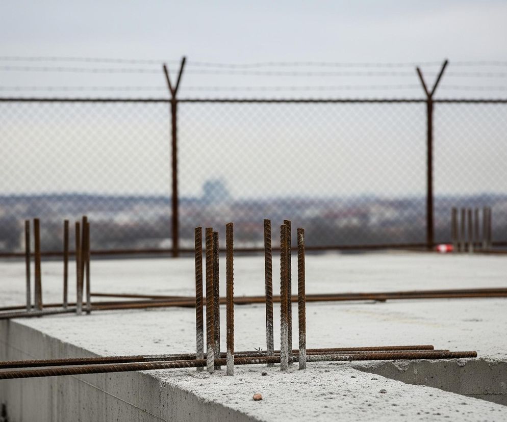Na pierwszym planie widoczny jest fragment betonowej konstrukcji budowlanej, z której wystają liczne pręty zbrojeniowe. Pręty są częściowo pokryte zaschniętym cementem, a ich powierzchnia ma rdzawy, chropowaty wygląd. Za betonową płaszczyzną, w oddali, rozciąga się metalowe ogrodzenie z siatki, zwieńczone drutem kolczastym rozpiętym na słupkach w kształcie litery V. Tło stanowi zamglony, szary krajobraz miejski pod pochmurnym niebem.