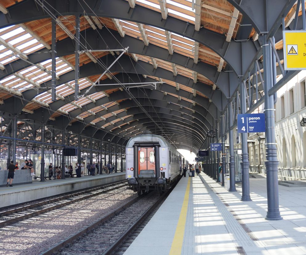 Peron na stacji kolejowej z pociągiem stojącym przy torze. Widać fragmenty zadaszenia, sieć trakcyjną oraz ludzi czekających lub przechodzących. Inwestycje w kolej to temat na Super Biznes.