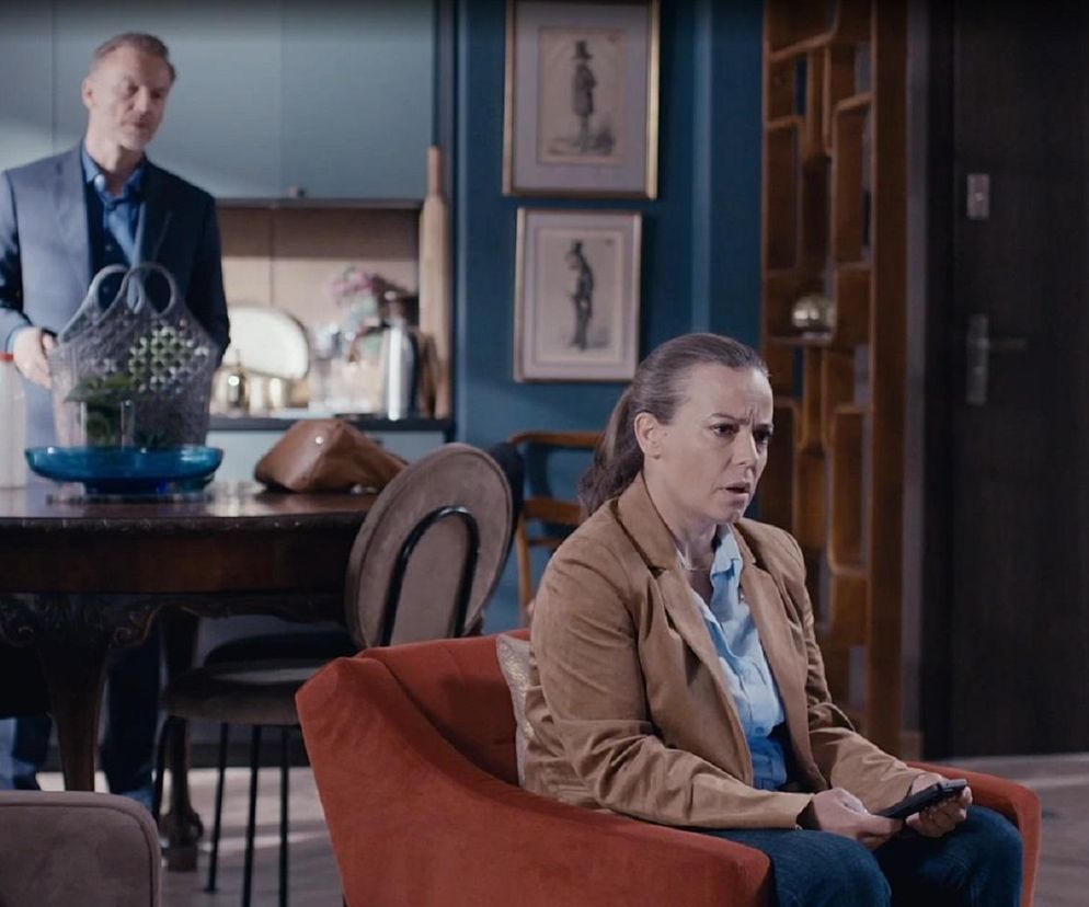 Magda (Anna Mucha) w brązowej marynarce siedzi na pomarańczowym fotelu z pilotem w ręku, zmartwiona. W tle Andrzej (Krystian Wieczorek) stoi przy stole, trzymając koszyk z zakupami. Scena ilustruje dylematy Magdy związane z powrotem do kancelarii w M jak miłość, o czym więcej przeczytasz na Super Seriale.