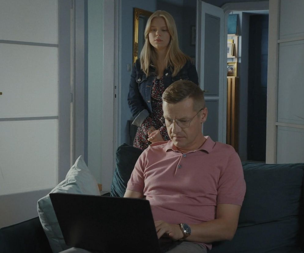 Piotrek Zduński (Marcin Mroczek) w różowej koszulce polo i okularach siedzi na kanapie, intensywnie pracując na laptopie. Za nim, w głębi pokoju, stoi jego córka Lenka (Olga Cybińska) w kwiecistej sukience i jeansowej kurtce, patrząc z zamyśleniem. Scena z serialu M jak miłość o problemach córki Zduńskich do przeczytania na Super Seriale.
