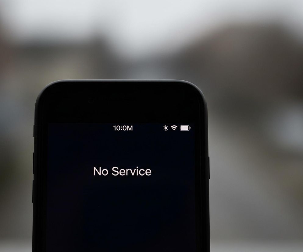 Czarny smartfon z zaokrąglonymi rogami, zajmujący dolną połowę kadru, wyświetla biały tekst No Service na czarnym tle. W górnej części ekranu, widoczny jest czas 10:0M po lewej stronie oraz ikony stanu baterii, Wi-Fi i Bluetooth po prawej. Tło za telefonem jest rozmyte, dominują odcienie szarości i brązu, sugerując plener lub wnętrze z oddalonymi obiektami.