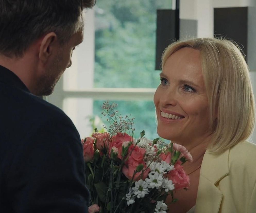 Uśmiechnięta Dorota (Iwona Rejzner) z jasnymi włosami trzyma bukiet różowych i białych kwiatów, patrząc na Bartka (Arkadiusz Smoleński), którego widać z tyłu. Scena z serialu M jak miłość przedstawia radosny moment Doroty i Bartka przed tragicznym wypadkiem Franki i Pawła, o czym przeczytasz na Super Seriale.