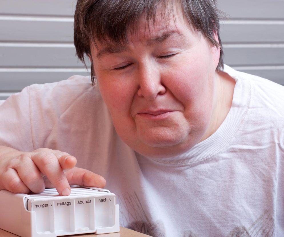 Kobieta z zamkniętymi oczami i zatroskaną miną, w białej koszulce, sięgająca palcem do pojemnika na leki z przegródkami morgens, mittags, abends, nachts. Obraz symbolizuje trudności w dostępie do świadczeń wspierających dla osób z niepełnosprawnościami, o czym czytaj na Poradnik Zdrowie.