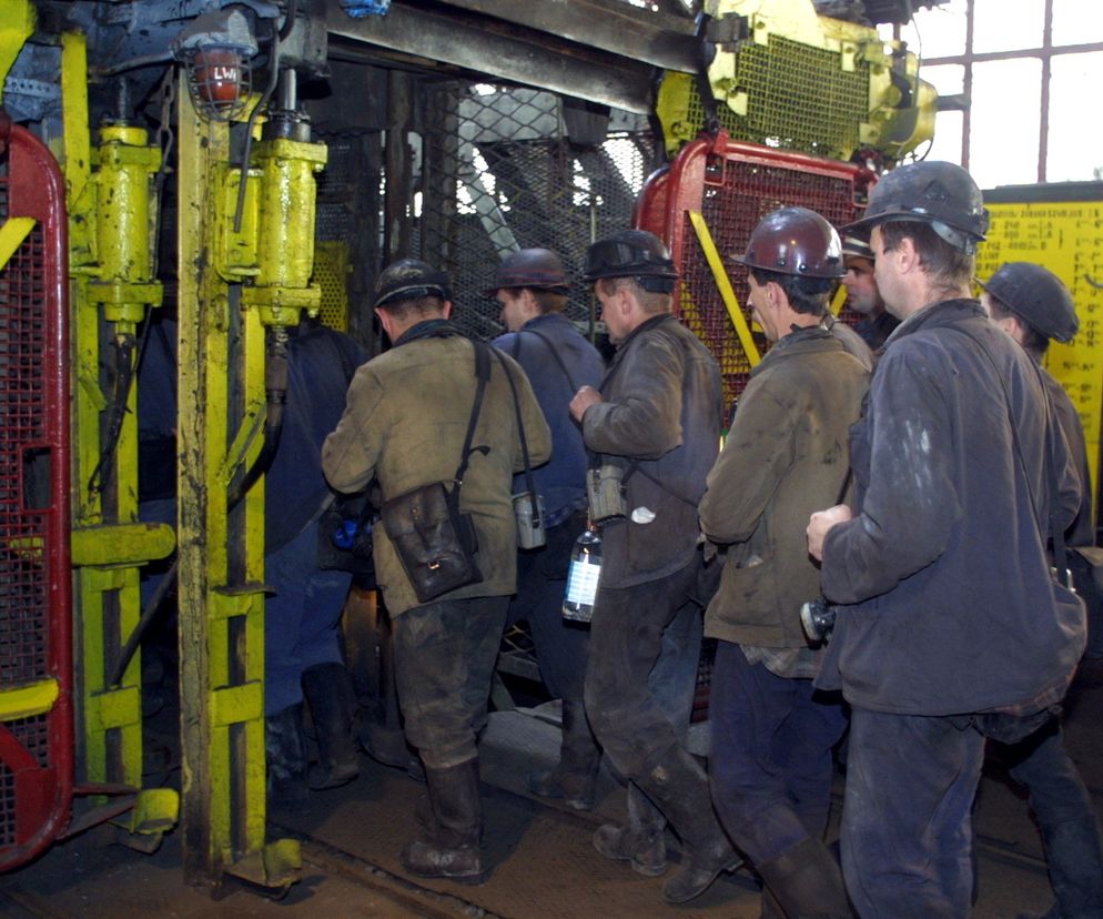 Grupa górników w kaskach i roboczych ubraniach wchodzi do szybu kopalnianego, przygotowując się do pracy. W tle widać stalowe konstrukcje i zabezpieczenia. O restrukturyzacji górnictwa można przeczytać na portalu Super Biznes.