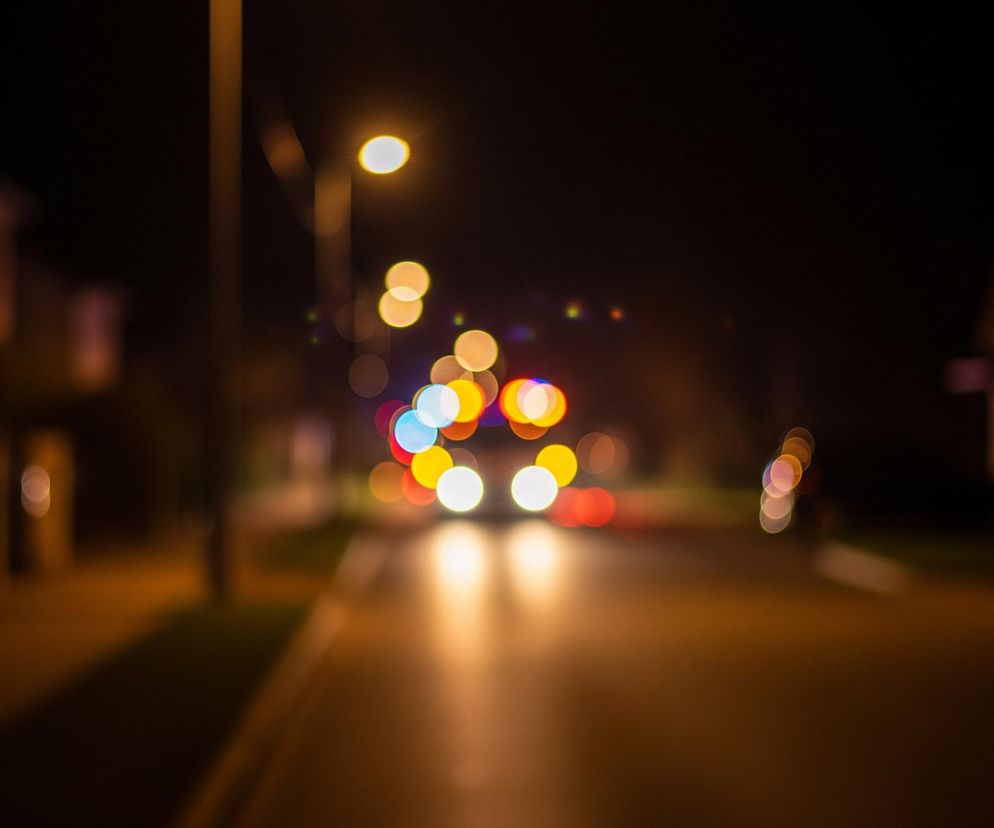 Ulica nocą rozciąga się w centralnej części kadru, z chodnikami po obu stronach. W tle widoczne są rozmyte światła samochodów i latarni ulicznych, tworzące kolorowe kręgi bokeh w odcieniach żółtego, białego, czerwonego i niebieskiego. Dwie jasne, białe plamy świetlne na dole centralnie wskazują na reflektory nadjeżdżającego pojazdu, oświetlając fragment asfaltu ciepłym, pomarańczowym blaskiem. Po lewej stronie widać niewyraźny obrys domu oraz słupa latarni, która emituje żółte światło.