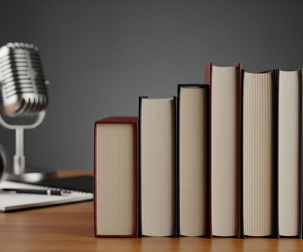 Na brązowym, błyszczącym blacie ułożonych jest siedem książek z widocznymi stronami, tworzących falę, z których dwie mają grzbiety w kolorze burgundowym, a pozostałe pięć w kolorze czarnym, rosnąc od lewej do prawej. Za książkami, po lewej stronie, stoi srebrny, stylizowany mikrofon retro, a obok niego leżą czarne słuchawki na otwartym notesie z długopisem. Tło stanowi jednolita, ciemnoszara ściana.