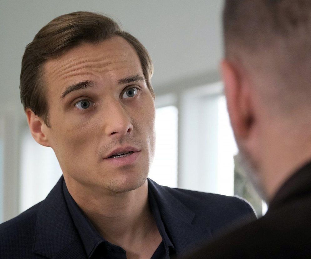 Zbliżenie na twarz Maurycego Popiela (Olek) z serialu M jak miłość, ożywiona ekspresja podczas rozmowy, z widocznym fragmentem pleców drugiej osoby. Scena nawiązuje do dramatycznego szantażu, z jakim Olek zmierzy się w odcinku 1879. Więcej o tym na Super Seriale.