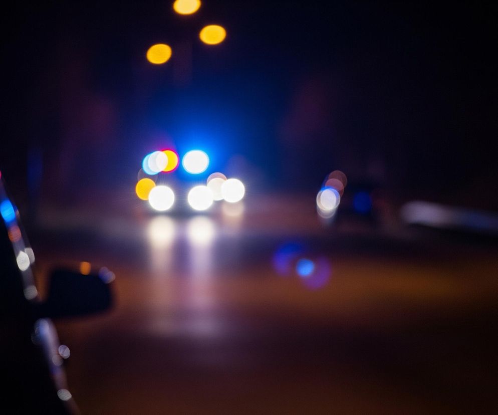 Rozmyty widok nocnej sceny, prawdopodobnie ulicy. Po lewej stronie, na pierwszym planie, widoczna jest niewyraźna część ciemnego samochodu, z odblaskami światła na jego powierzchni i lusterku bocznym. W tle, na ciemnym niebie, jaśnieją trzy żółte, okrągłe światła. Poniżej, na środku obrazu, znajduje się samochód z włączonymi, rozmazanymi światłami: dwie jasne białe kule to reflektory, a nad nimi migoczą niebieskie i czerwone światła, sugerujące syreny policyjne lub ratunkowe. Cała scena jest ciemna, z pojedynczymi, rozmyślanymi plamami światła w różnych kolorach.