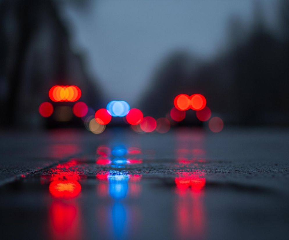 Mokry asfalt w pierwszym planie odbija jaskrawe, rozmyte światła samochodów. Na drodze widać liczne okrągłe, czerwone i niebieskie refleksy, sugerujące obecność świateł pozycyjnych lub sygnalizacyjnych, odbijające się od wilgotnej nawierzchni. W tle, powyżej horyzontu drogi, widoczne są podobne, rozmyte światła pojazdów: dominują okrągłe czerwone światła, prawdopodobnie tylne, oraz jedno niebieskie w centrum, mogące wskazywać na samochód uprzywilejowany. Tło stanowią ciemne, niewyraźne kształty drzew i budynków, zlewające się z ponurym, szarym niebem.