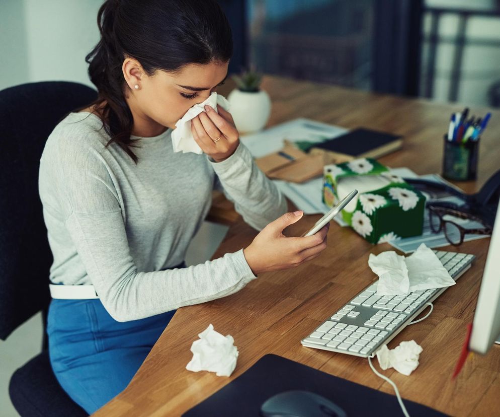 Kobieta w jasnej bluzce i niebieskich spodniach siedzi przy biurku, trzymając w lewej ręce smartfon, a prawą dłoń z chusteczką przy nosie, sprawiając wrażenie chorej i przemęczonej. Wokół niej leżą zużyte chusteczki oraz pudełko z chusteczkami, co wskazuje na jej złe samopoczucie. Temat absencji chorobowej i pracy mimo choroby można zgłębić na portalu Poradnik Zdrowie.