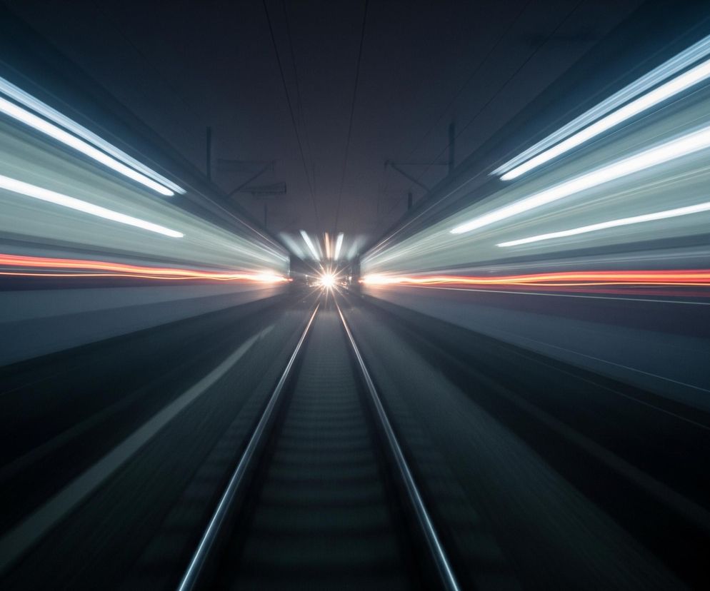 Ciemny tunel, przez który biegnie para torów kolejowych, znikających w oddali w punkcie zbieżności. Dynamiczne smugi światła, głównie białe i jasnoniebieskie, rozciągają się horyzontalnie po obu stronach kadru, tworząc wrażenie dużej prędkości. W centrum, na końcu torów, widoczne jest jasne, rozproszone źródło światła, otoczone promieniami. Pod smugami światła na bocznych ścianach tunelu biegną cienkie, pomarańczowo-czerwone linie światła.