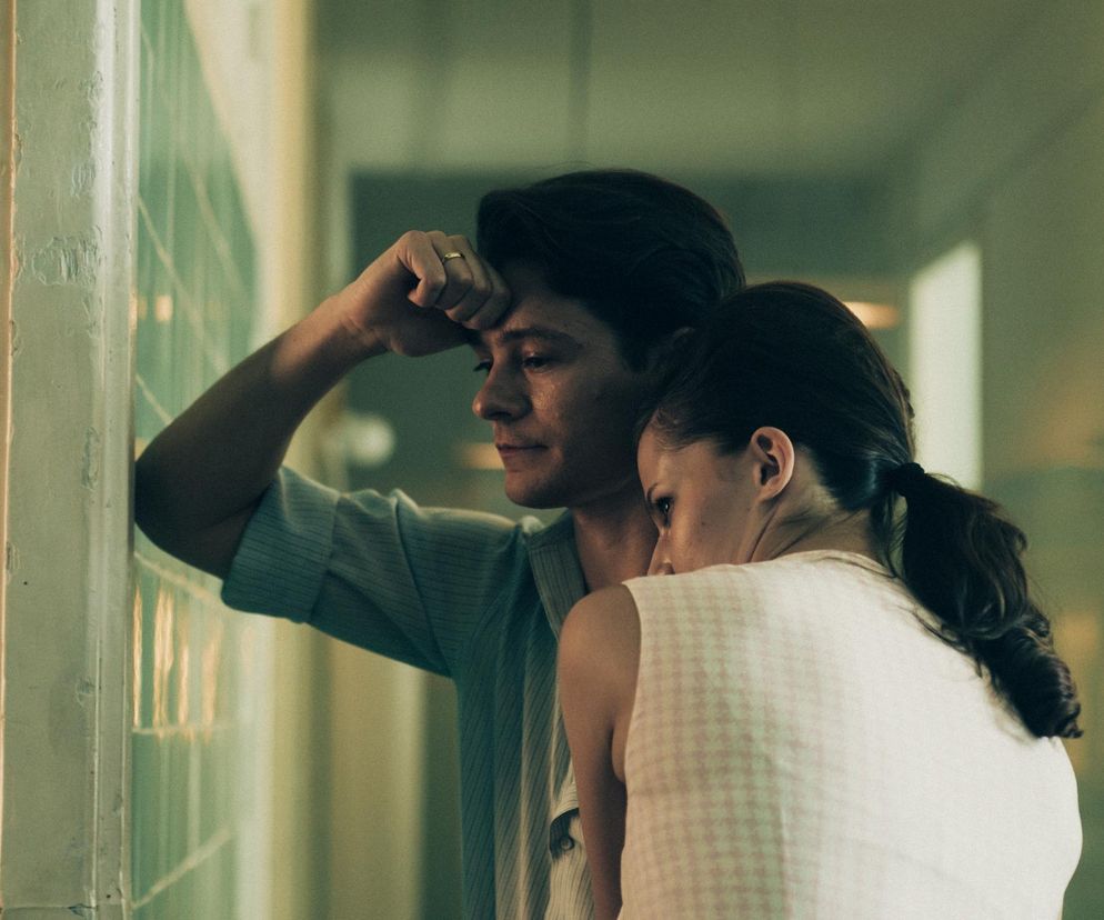 Mężczyzna z obrączką na palcu opiera głowę na dłoni, patrząc w bok, obok niego stoi kobieta z włosami związanymi w kucyk, opierająca głowę o jego ramię. Oboje wyglądają na zasmuconych w szpitalnym otoczeniu. Zdjęcie przedstawia Tomasza Ziętka i Agatę Łabno w roli Igora i Anny Bielików z serialu Czarna śmierć. Więcej o tym na Super Seriale.