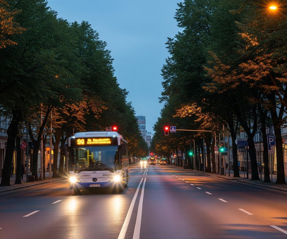 Autobus miejski z włączonymi światłami mijania i kierunkowskazem skrętu w lewo jedzie środkowym pasem ulicy, w tle widoczne są światła innych pojazdów. Ulica jest dwupasmowa w każdym kierunku, oddzielona podwójną ciągłą linią, a po obu stronach znajduje się chodnik z rzędem drzew. Drzewa oraz część jezdni są oświetlone ciepłym, pomarańczowym światłem latarni ulicznych, natomiast sygnalizacja świetlna w oddali świeci na czerwono i zielono. Niebo jest granatowe, wskazując na zmierzch lub wczesny ranek.