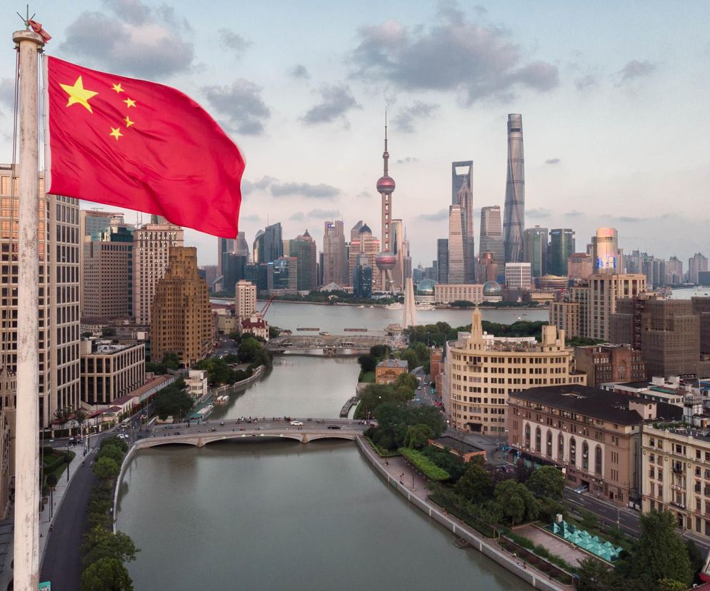 Flaga Chin powiewa nad panoramą Szanghaju, z rzeką w centrum oraz mostami, ulicami i nowoczesnymi wieżowcami w tle. Widoczna jest też Wieża Perły Orientalnej. To kontekst do artykułu o ustawie Lex Huawei i relacjach z Chinami na portalu Super Biznes.