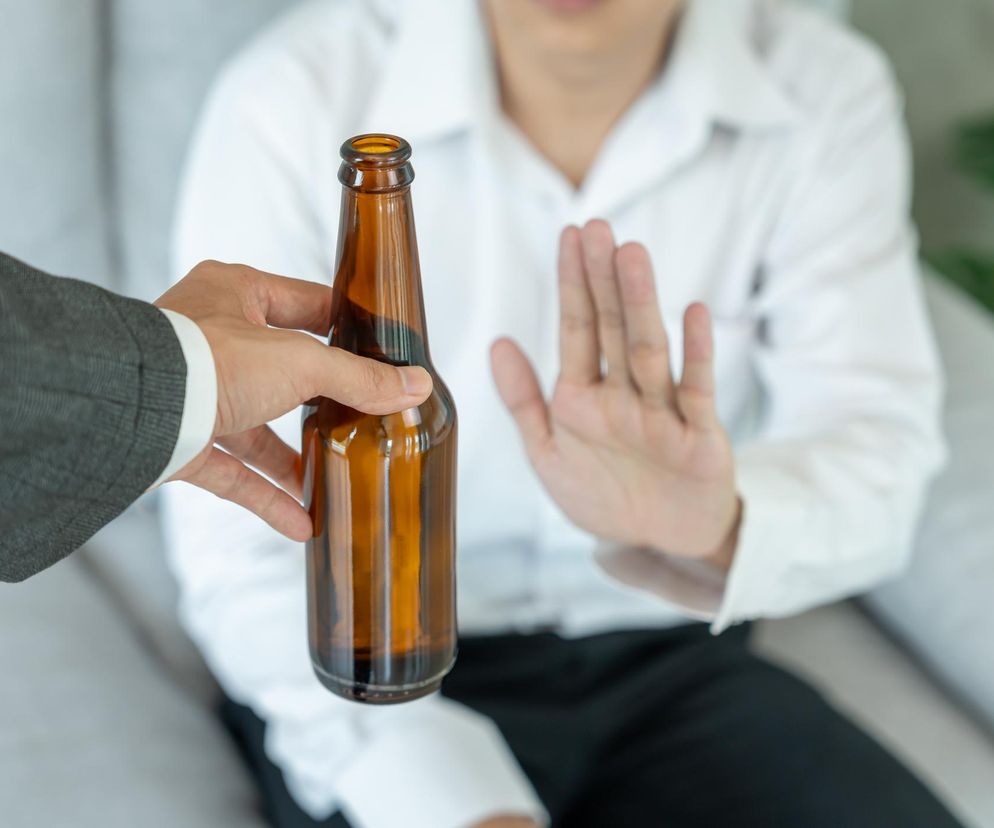 Osoba w białej koszuli odmawia przyjęcia butelki alkoholu, trzymając otwartą dłoń w geście stop, co symbolizuje rosnący trend ograniczania spożycia alkoholu wśród Polaków, o czym przeczytasz na Poradnik Zdrowie.