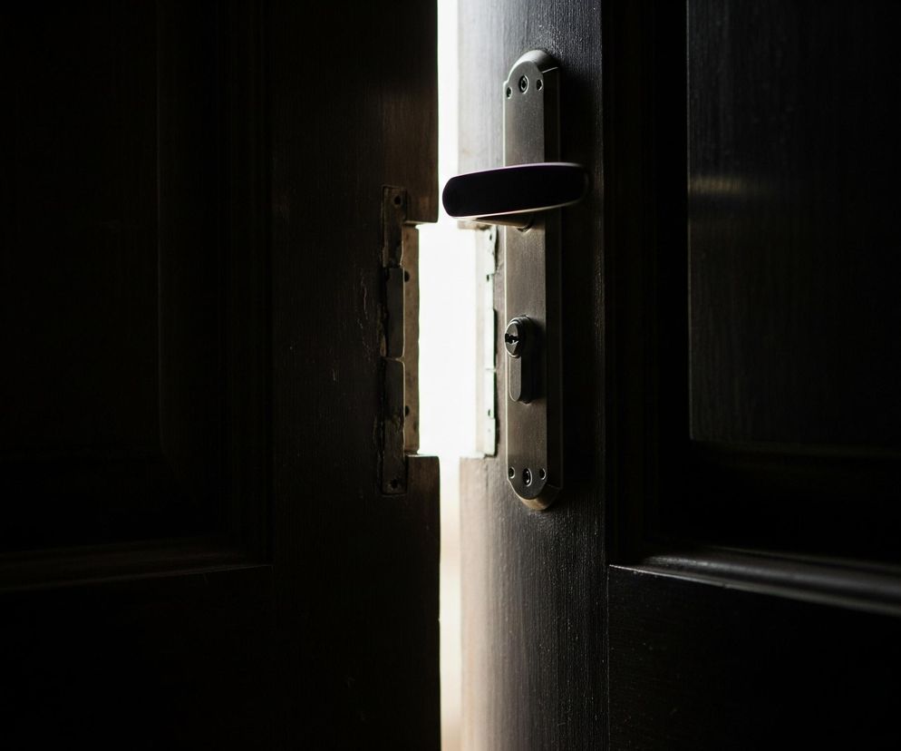 Ciemne drewniane drzwi są uchylone, tworząc wąską, pionową szczelinę jasnego światła pośrodku kadru. Po prawej stronie szczeliny widoczna jest ciemna klamka z wbudowanym zamkiem, osadzona w masywnej drewnianej płycie drzwiowej. Reszta obrazu, po lewej i prawej stronie centralnego światła, jest niemal całkowicie czarna, z ledwo widocznymi zarysami innych elementów drzwiowych, co podkreśla kontrast między światłem a cieniem.