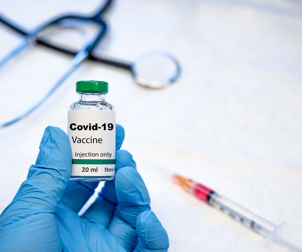 Ręka w niebieskiej rękawiczce trzyma fiolkę ze szczepionką Covid-19, z napisem Injection only, 20 ml. W tle widać stetoskop i strzykawkę z różową substancją, symbolizujące badania nad odpornością po szczepieniu, o czym więcej przeczytasz na Poradnik Zdrowie.