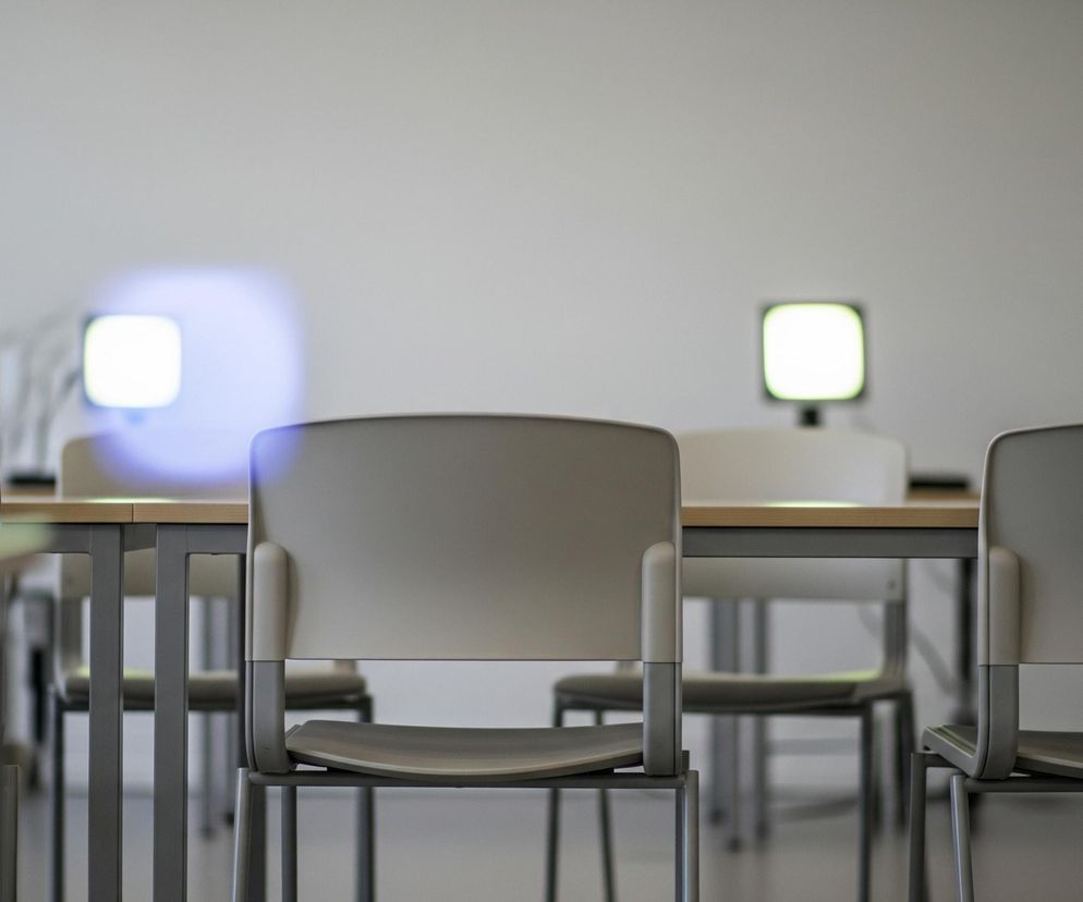 W centrum obrazu widoczne jest beżowo-szare krzesło z zaokrąglonymi oparciem i siedziskiem, stojące na tle jasnej, szarej ściany. Przed krzesłem znajduje się drewniany blat stołu, podtrzymywany przez ciemnoszare, metalowe nogi. Po lewej i prawej stronie tego głównego krzesła, częściowo zakryte, widoczne są podobne stoły i krzesła, tworząc szereg mebli. W oddali, na jasnej ścianie, widać dwa rozmyte, prostokątne obszary światła: jeden biało-niebieskawy po lewej i jeden biało-zielonkawy po prawej.