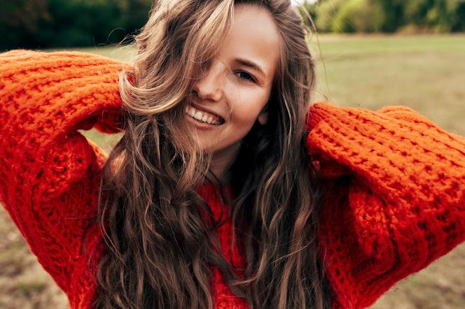 Outdoor portrait of a smiling young woman wearing a knitted orange sweater posing on nature background. The beautiful female has a joyful expression, resting in the park. Włosy sweter