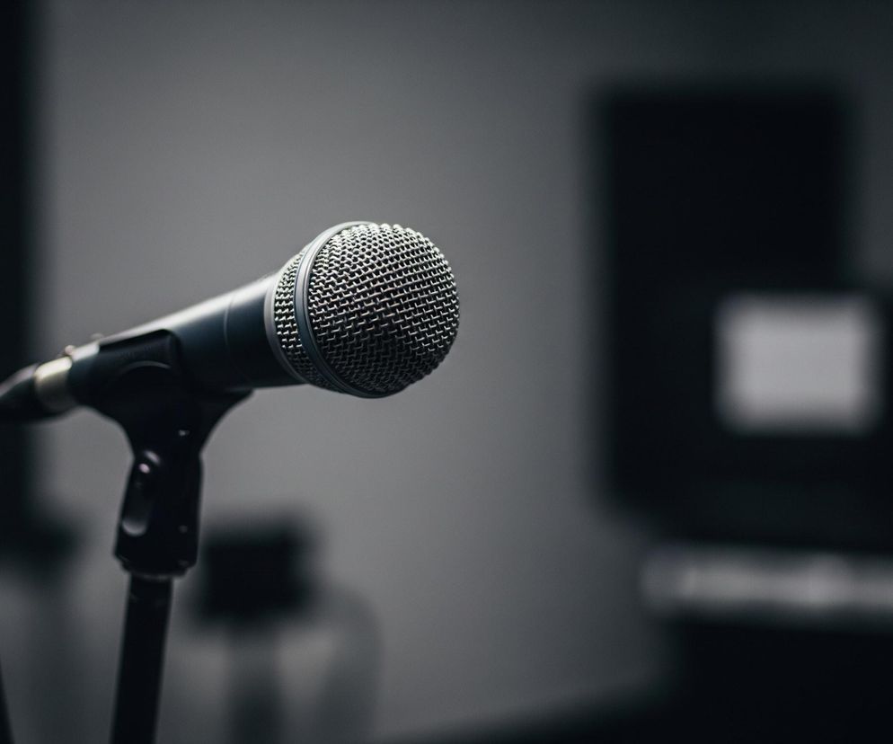 Srebrny mikrofon z cylindryczną siateczką na górze znajduje się w centrum kadru, lekko skierowany w prawo. Mikrofon zamocowany jest na czarnym statywie, który znika w dolnej części obrazu. Tło jest rozmyte, utrzymane w ciemnych, szarych odcieniach, z niewyraźnymi, jaśniejszymi i ciemniejszymi plamami. Całość jest słabo oświetlona, co nadaje scenie stonowany charakter.