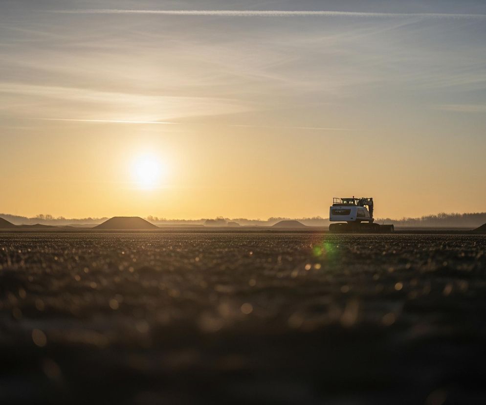 Na rozległym, otwartym terenie, przypominającym świeżo zaoraną ziemię, znajduje się koparka z wyraźnie widocznym napisem LIEBHERR na boku, umieszczona w centralnej części obrazu. Słońce, intensywnie jasnożółte, świeci nisko nad horyzontem po lewej stronie kadru, rzucając długie cienie i tworząc silne refleksy świetlne na ziemi. Niebo jest rozświetlone odcieniem pomarańczowo-żółtym u dołu, przechodzącym w jaśniejszy błękit ku górze, z widocznymi delikatnymi smugami chmur. Na horyzoncie, w tle, widoczne są niskie, zaokrąglone wzgórza ziemi oraz linia lasu, z niewyraźnymi sylwetkami drzew.