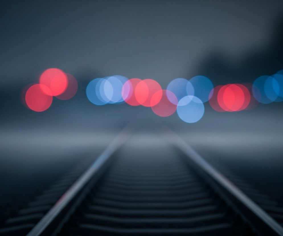 Na pierwszym planie widoczne są dwie równoległe szyny kolejowe z podkładami, prowadzące wprost do centrum obrazu, stając się coraz mniej wyraźne w oddali. Tło rozmyte jest w efektownym bokeh, ukazując szereg okrągłych świateł w kolorach czerwonym i niebieskim. Cała scena utrzymana jest w chłodnej tonacji szarości i błękitu, co nadaje jej nieco tajemniczy i nastrojowy charakter.