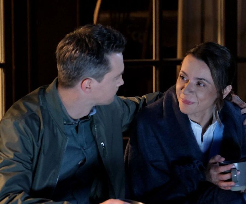 Mężczyzna w zielonej kurtce i kobieta w granatowym szlafroku trzymająca kubek, siedzą blisko siebie na zewnątrz budynku. Mężczyzna patrzy na kobietę, która uśmiecha się i patrzy w bok. Scena romantycznego wieczoru z serialu M jak miłość. Więcej o tym odcinku przeczytasz na Super Seriale.