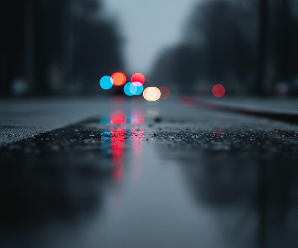 Mokry, asfaltowy chodnik lub ulica znajduje się na pierwszym planie, z drobnymi kroplami wody widocznymi na jego powierzchni. Przed nim biegnie niewyraźny pas ciemniejszego materiału. Na drugim planie, w centrum, widoczne są rozmyte, okrągłe światła samochodów: cztery czerwone, dwa jasnoniebieskie i jeden kremowy, z ich odbiciami w wodzie na chodniku. Tło jest całkowicie rozmyte, szare i ciemne, z zarysami drzew i budynków po obu stronach drogi.