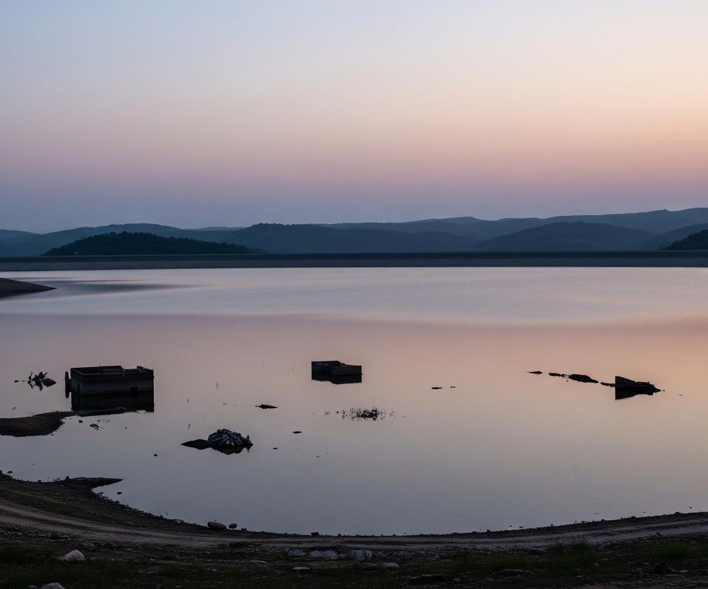 Centralnym elementem obrazu jest duży zbiornik wodny, którego powierzchnia odbija pastelowe kolory wieczornego nieba – od jasnego błękitu i fioletu w górnej części po ciepłe odcienie pomarańczy i różu po prawej stronie. Na pierwszym planie, po lewej stronie, widoczne jest nierówne, trawiaste i kamieniste zbocze, z którego schodzi ścieżka gruntowa, odsłaniając brzeg zbiornika. W wodzie, w różnych odległościach od brzegu, znajdują się zanurzone konstrukcje przypominające ruiny budynków, a także fragmenty drzew i inne ciemne obiekty, z których niektóre częściowo wystają ponad powierzchnię. Horyzont wyznacza pasmo ciemnych, zalesionych wzgórz, za którymi rozciąga się jaśniejsze niebo.