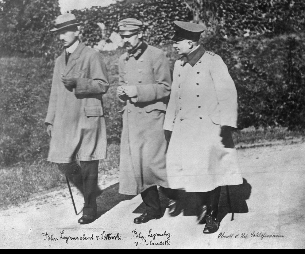 Józef Piłsudski, z wąsem, w legionowym płaszczu i rogatywce, maszeruje obok dwóch mężczyzn, prawdopodobnie oficerów niemieckich, podczas internowania w Magdeburgu. Historyczne ujęcie z czasów niewoli Piłsudskiego, o czym przeczytasz więcej na Super Biznes.