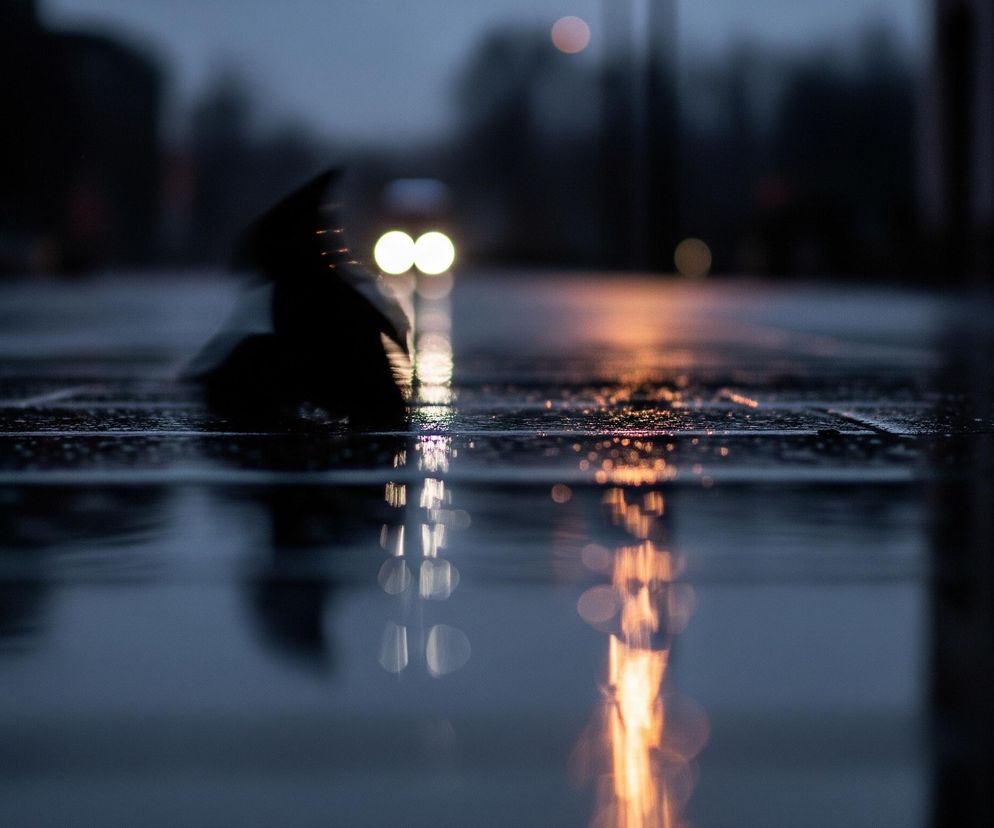 Na pierwszym planie widoczna jest mokra nawierzchnia, prawdopodobnie asfalt lub bruk, z licznymi odbiciami świetlnymi i drobnymi kroplami wody. W tle, rozmyte i nieostre, majaczą dwa jasne, koliste światła, przypominające reflektory samochodu, które odbijają się w kałuży, tworząc długie, pionowe smugi światła na powierzchni. Po prawej stronie, nieco dalej, widoczne jest pomarańczowe światło, również odbijające się w wodzie, co sugeruje obecność innego źródła światła miejskiego. Ciemna, zamazana sylwetka z lewej strony, być może człowieka z parasolem, porusza się w kierunku świateł samochodu. Całość jest utrzymana w ciemnych, chłodnych barwach, z dominacją granatu i czerni, przełamywanych jaskrawymi, ciepłymi światłami.