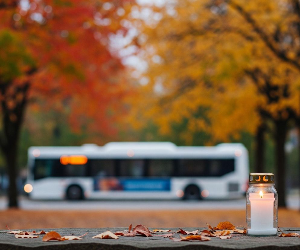 Na pierwszym planie, po prawej stronie, znajduje się zapalony znicz w przezroczystym szklanym naczyniu z metalową, złotawą nakrętką, na której widnieją czarne otwory. Znicz stoi na kamiennym murku lub parapecie, pokrytym licznymi, suchymi, jesiennymi liśćmi w odcieniach brązu, pomarańczu i żółci. W tle widać rozmyte, jesienne drzewa o liściach w intensywnych barwach czerwieni, pomarańczu i żółci, a między nimi, również nieostry, biały autobus z widocznym, pomarańczowym wyświetlaczem i niebieskimi elementami reklamy na boku.