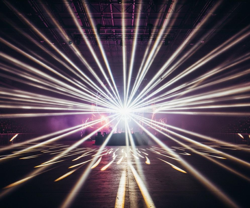 Centralny punkt obrazu to scena oświetlona silnym, białym światłem, z której rozchodzą się liczne promienie świetlne, tworząc efekt rozchodzącej się gwiazdy. Promienie te, w odcieniach bieli i jasnego złota, przecinają ciemną przestrzeń, oświetlając fragmenty podłogi w przedniej części obrazu. Tło sceny wypełnia delikatna, fioletowo-różowa mgła, a na skrajach kadru widoczne są fragmenty ciemnych trybun z rozproszonymi, małymi, kolorowymi światłami.