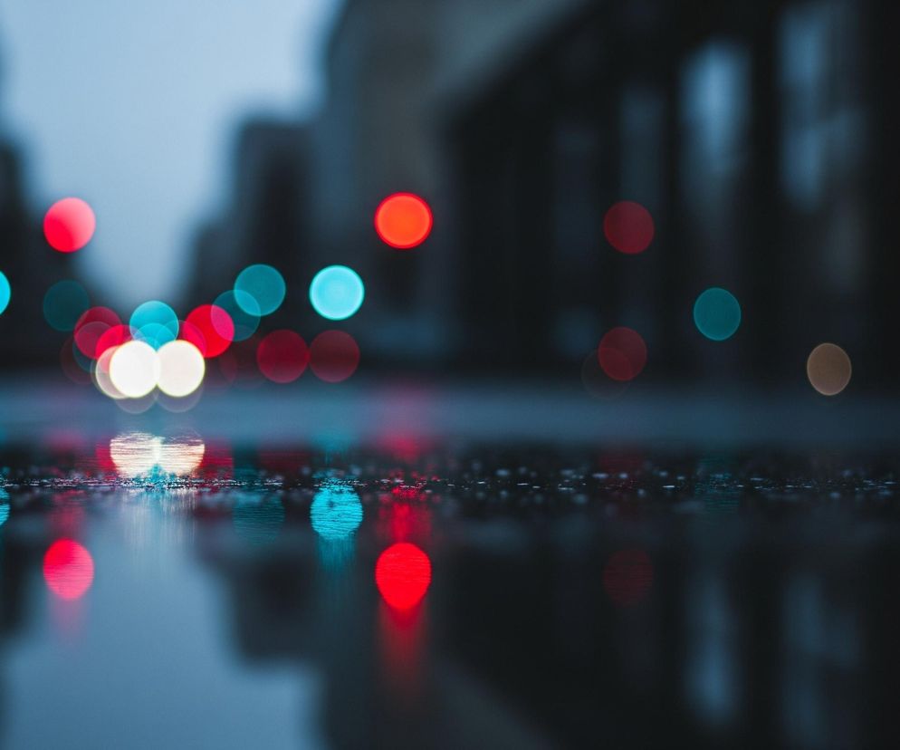 Przedstawiono rozmyty widok miejskiej ulicy o zmierzchu lub w nocy, z licznymi, nieostrymi światłami pojazdów i sygnalizacji świetlnej. Powierzchnia drogi jest mokra, odbijając barwne światła. Ulica wydaje się pusta, a tło stanowią niewyraźne zarysy budynków.