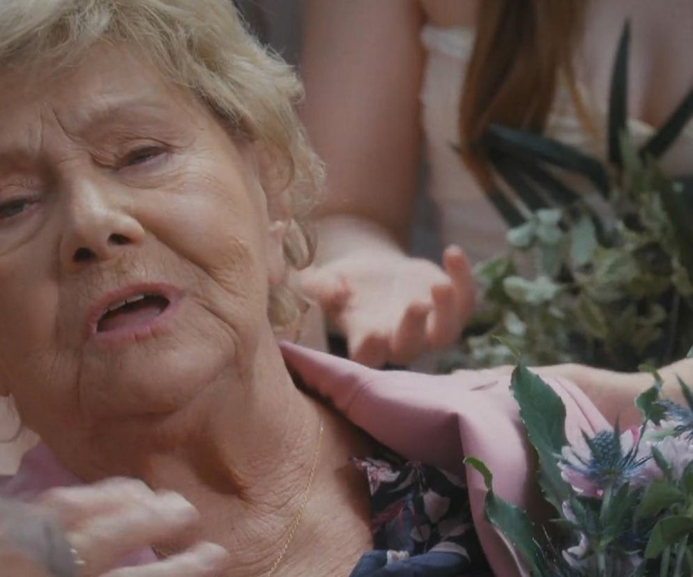 Barbara Mostowiak z serialu M jak miłość z bladą twarzą, udająca omdlenie na ślubie Kamy i Marcina, leży wsparta przez inną osobę, trzymając w ręku bukiet. Zdjęcie ilustruje scenę z serialu, a więcej szczegółów znajdziesz na Super Seriale.