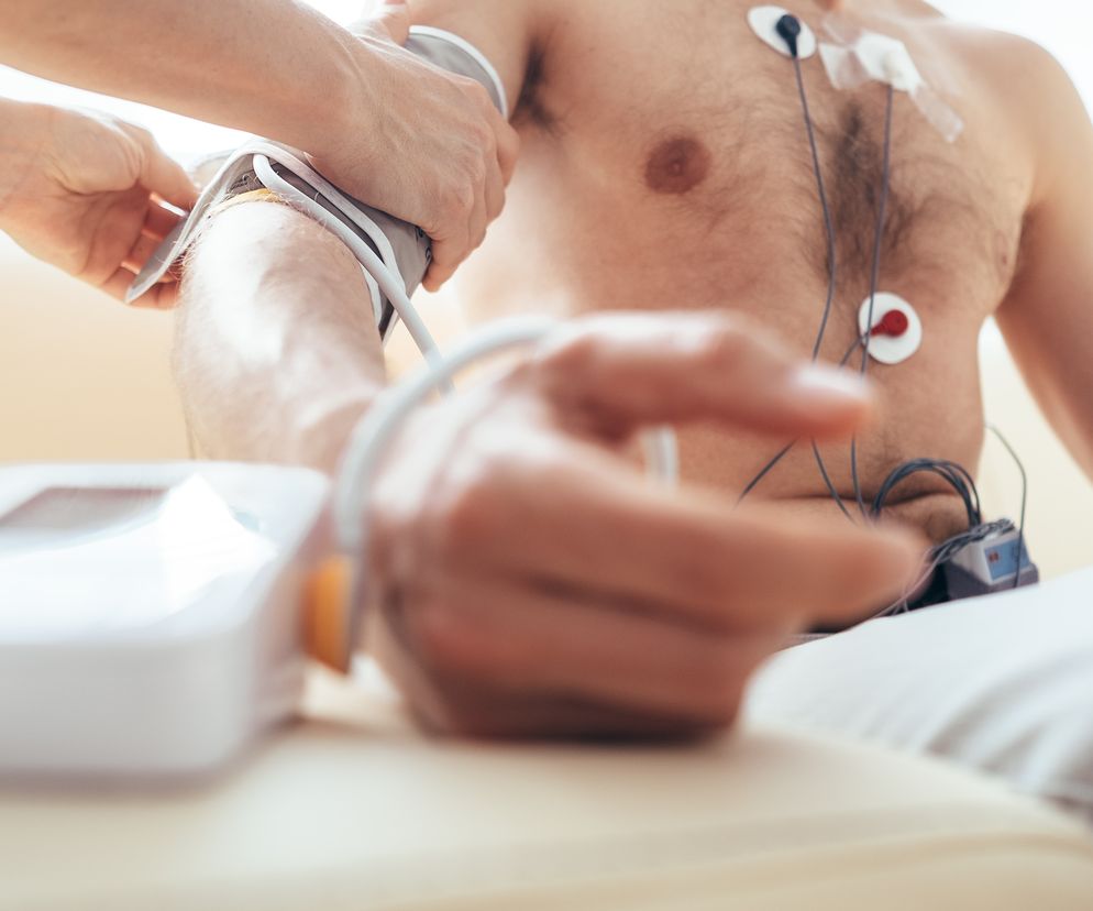 Mężczyzna z podłączonym Holterem EKG i elektrodami na klatce piersiowej, któremu mierzone jest ciśnienie krwi na ramieniu przez niewidoczną osobę. Blisko widoczne urządzenie do pomiaru ciśnienia. Więcej o badaniu metodą Holtera przeczytasz na Poradnik Zdrowie.