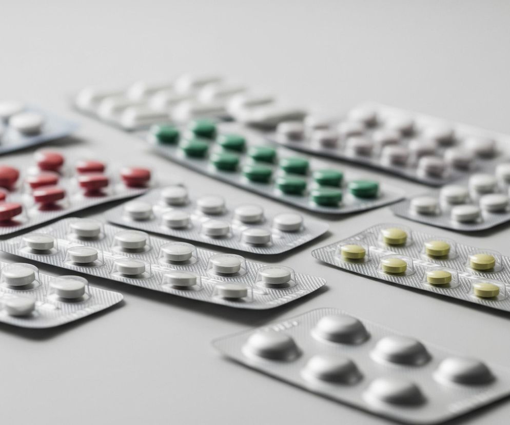 Na jasnoszarym tle rozłożonych jest dziewięć opakowań listkowych z tabletkami. Większość opakowań, zawierających białe, czerwone i jasnozielone pigułki, jest częściowo wypełniona, a każde opakowanie ma kształt prostokąta z aluminiowym spodem i przezroczystymi, wypukłymi blistrami. Kilka opakowań widocznych jest w całości, leżąc obok siebie, podczas gdy pozostałe są częściowo zamazane, z widocznym jedynie fragmentem ich kształtu.