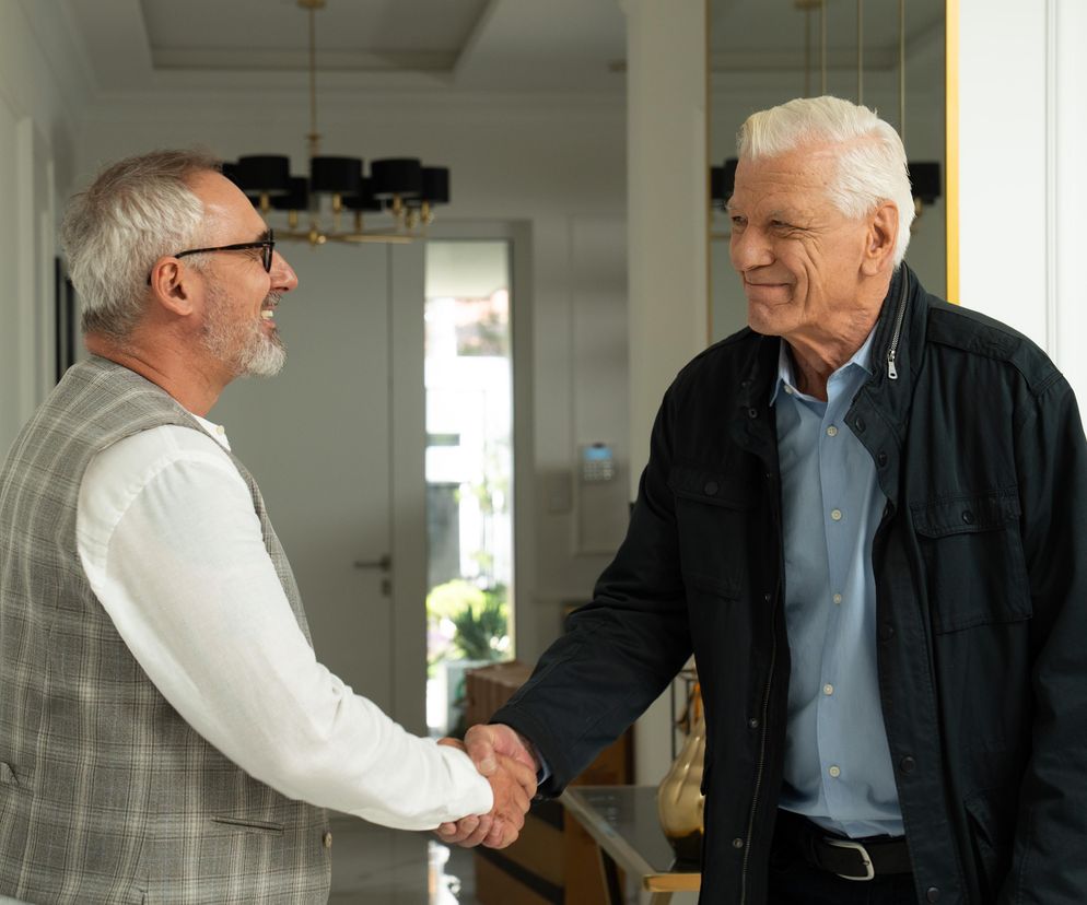 Uśmiechnięci Adam Werner (Jacek Kopczyński) i pan Mirek (Marcin Sosnowski) z serialu M jak miłość uściskiem dłoni witają się w luksusowej posiadłości. Na zdjęciu widać eleganckie wnętrze, jasne ściany i duży złoty żyrandol w tle, co zapowiada zmiany w Super Seriale.