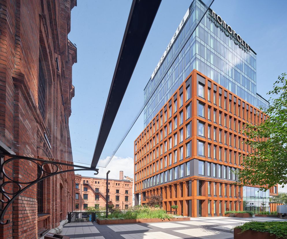 Nowoczesny budynek biurowy Monopolis II z elementami szkła i stali Corten obok zabytkowych, ceglanych zabudowań. W tle widać starsze, przemysłowe budynki, a na pierwszym planie taras z zielenią. Więcej o projekcie Monopolis II przeczytasz na portalu Architektura Murator Plus.